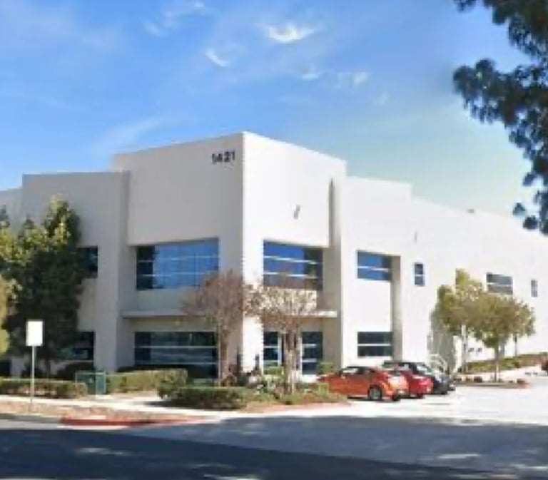 Modern white commercial building with blue-tinted windows, landscaping, and red vehicles parked in front under a clear sky