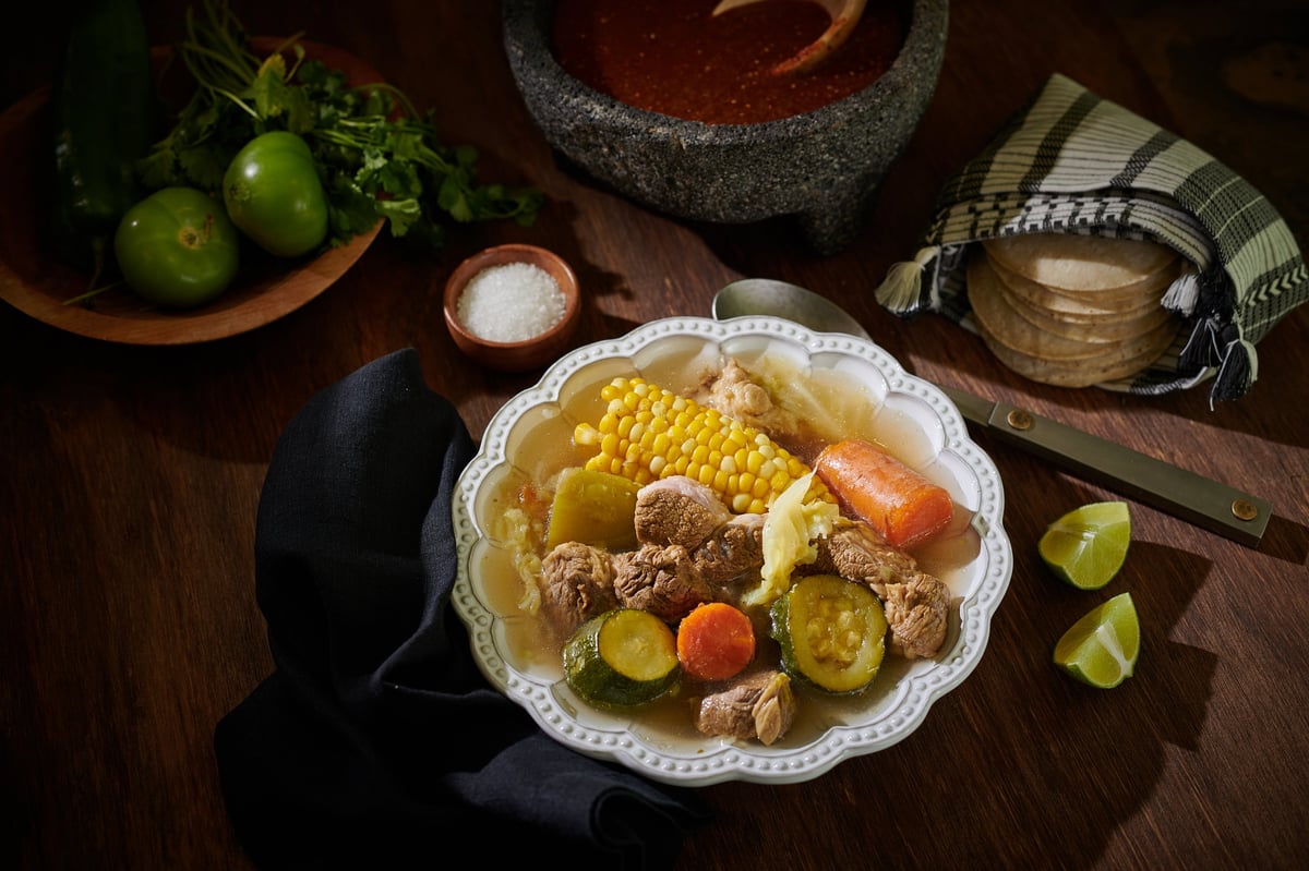Overhead view of a rustic plated dish with chicken, corn, carrots, and potatoes surrounded by fresh limes, salt, and cooking ingredients on a dark wooden table