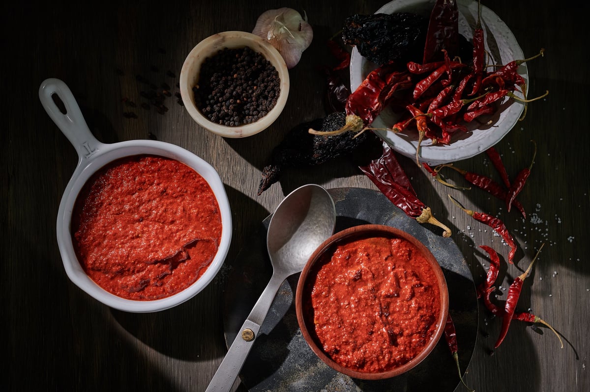 Overhead view of ground red chili powder in white bowls and pans with whole dried red chilis and black peppercorns on dark wood
