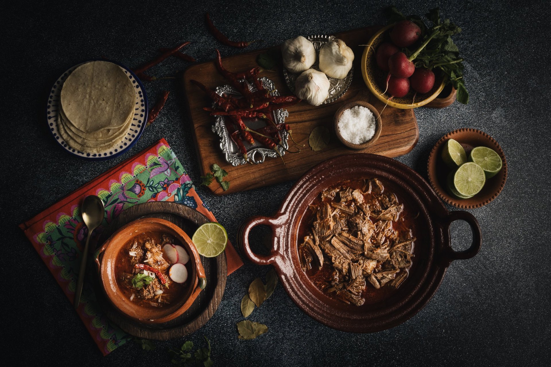 Overhead view of Mexican cooking ingredients and dishes including tortillas, two clay bowls with prepared food, raw ingredients like garlic and lime, arranged on a dark textured surface